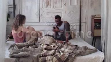 年轻男子在移动胶卷相机上拍摄穿睡衣的女人。 多种族的夫妇早上坐在床上。